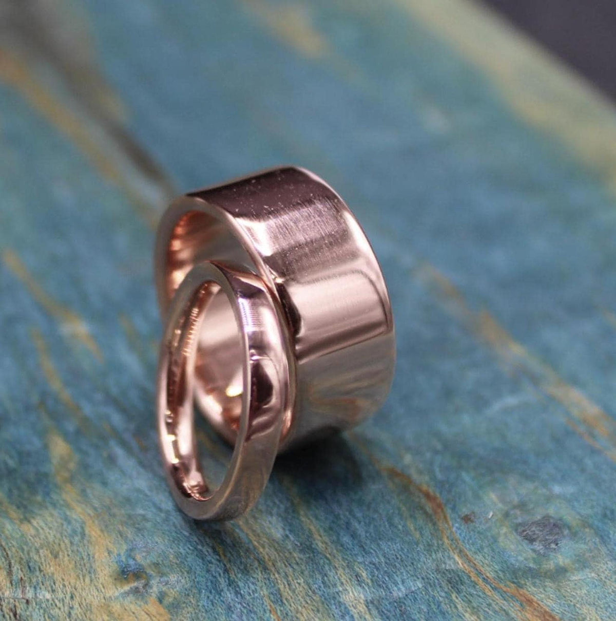 A thin, shiny, rose gold-colored ring leans against a wider, matching ring on a blue-green distressed wooden surface.