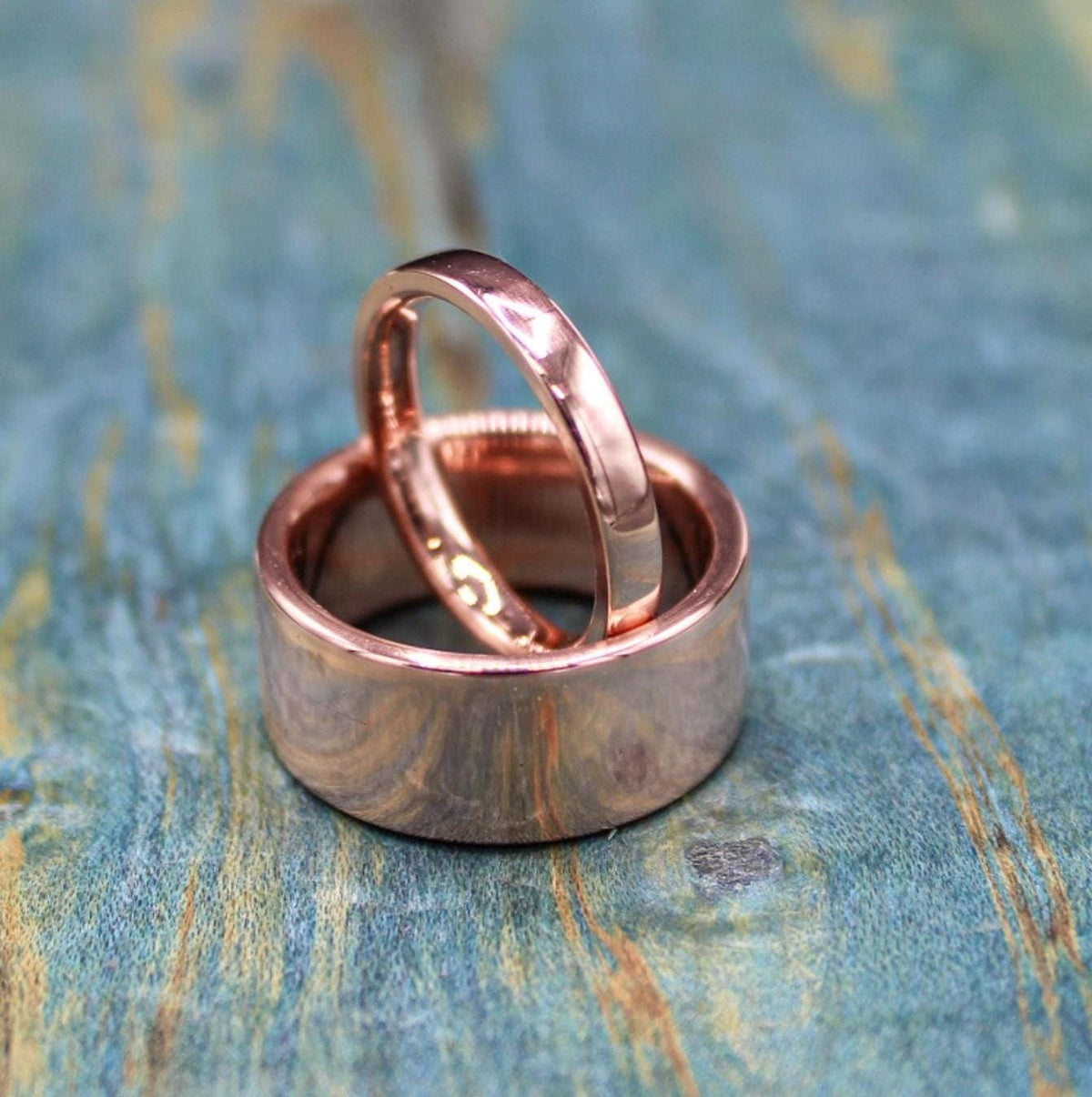 A narrow, shiny, rose gold-colored ring rests inside a wider, matching ring on a blue-green distressed wooden surface.