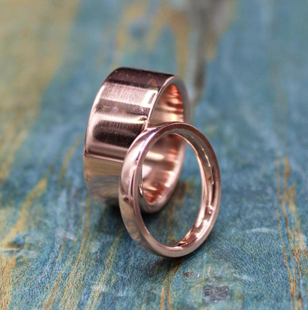 A wide, shiny, rose gold-colored ring is positioned behind a narrower, matching ring on a blue-green distressed wooden surface.