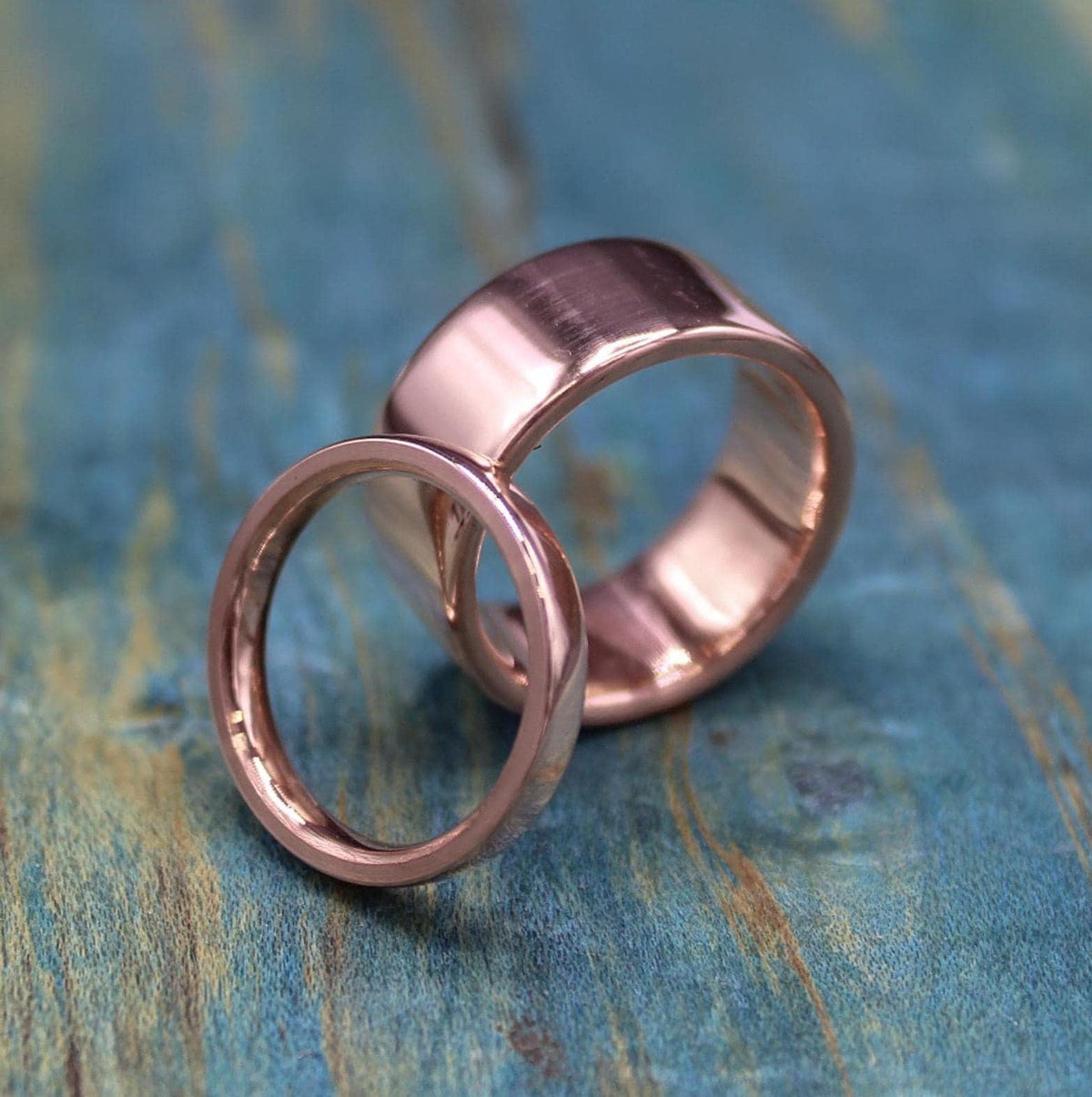 Two shiny, rose gold-colored rings, one thin and one wide, are angled on a blue-green distressed wooden surface.