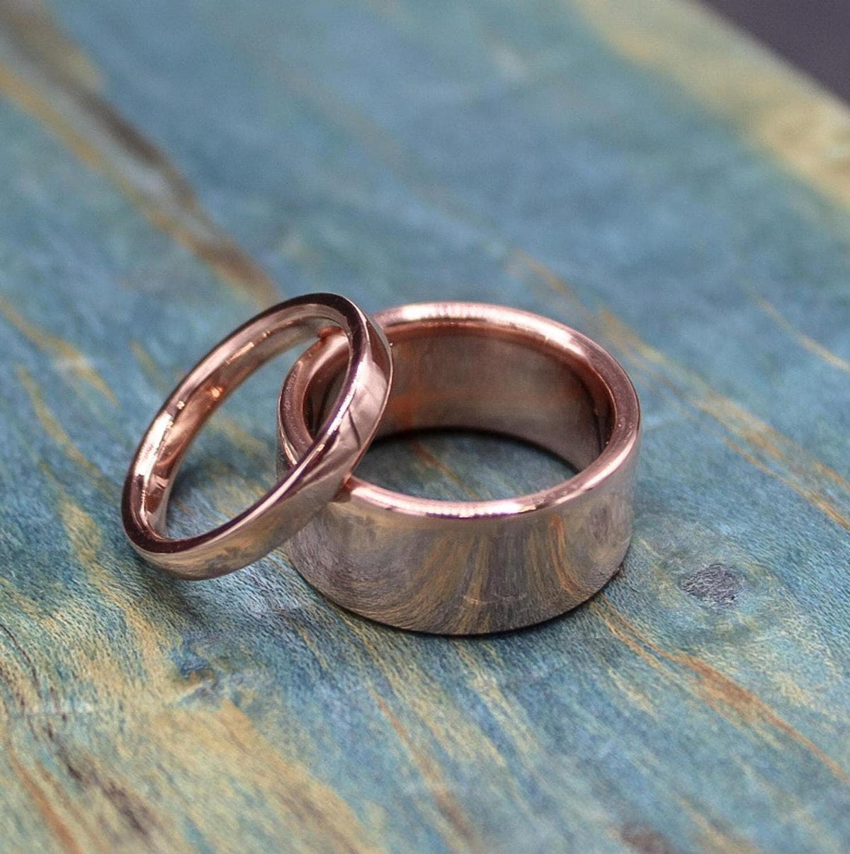 Two shiny, rose gold-colored rings, one narrow and one broad, are placed side-by-side on a blue-green distressed wooden surface.