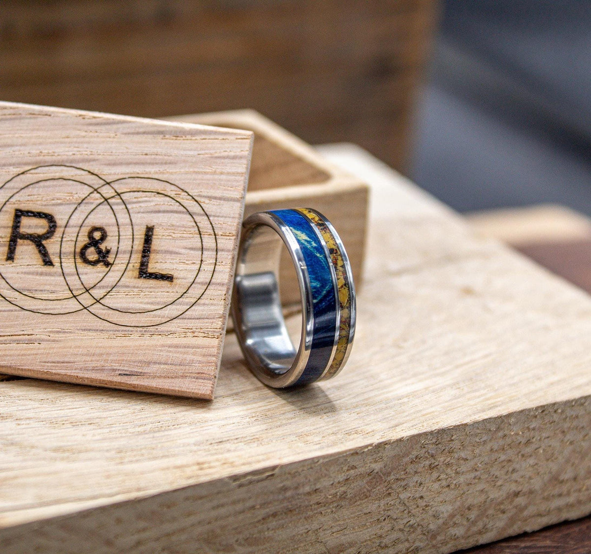 A silver-colored ring with a blue and yellow patterned inlay sits on a light wooden surface in front of an open wooden box with an "R & L" logo.