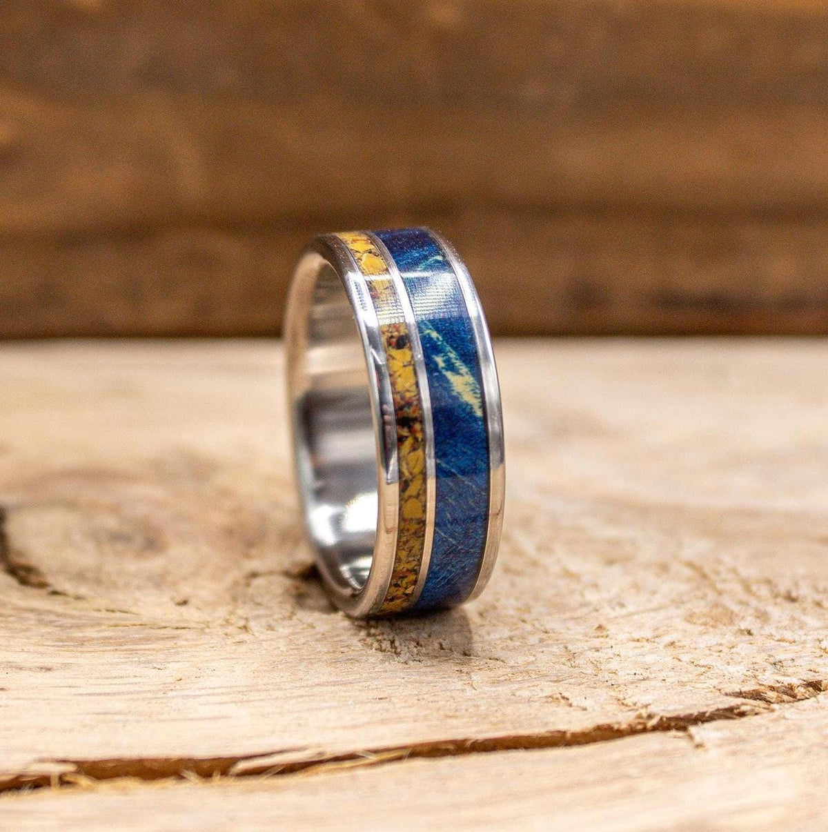 A silver-colored ring with a central yellow and blue patterned band is shown from a slight angle on a light wooden surface.