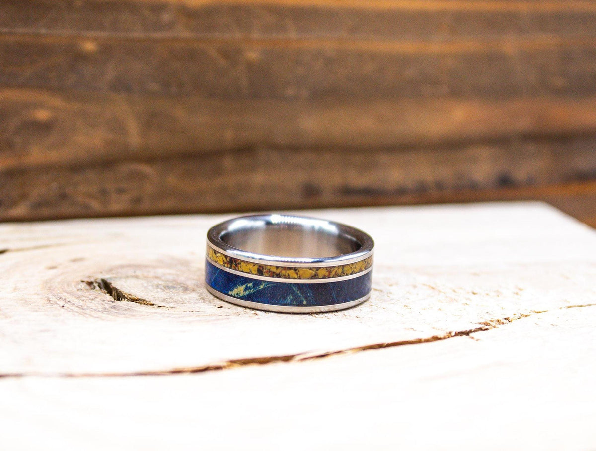 A silver-colored ring with a yellow and blue patterned inlay is centered on a light wooden surface, with a blurred wooden background.