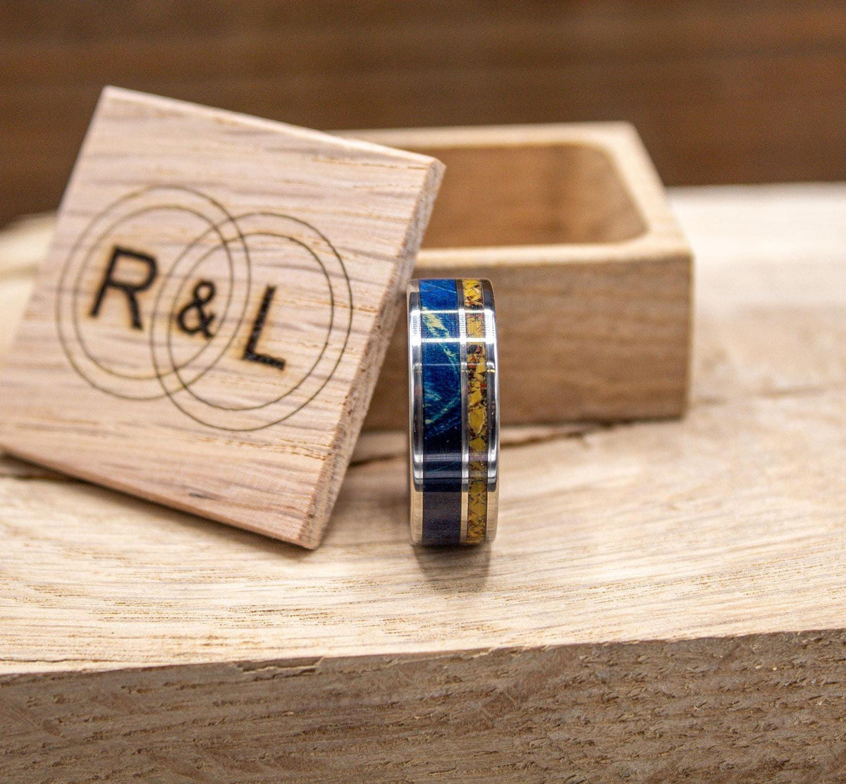 A silver-colored ring with a yellow and blue inlay is positioned next to an open wooden box with an "R & L" logo on a light wooden surface.