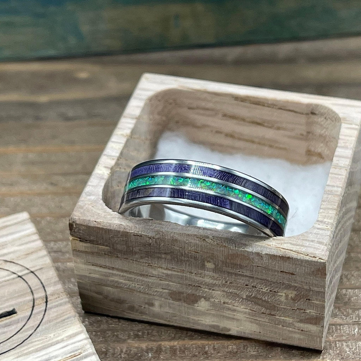 A silver ring with purple wood and green opal inlay is displayed in an open wooden box with a white cushion.