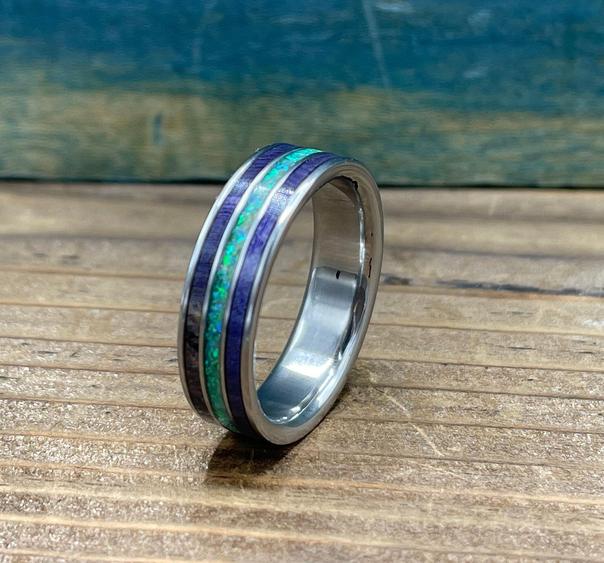 A silver ring with purple wood and green opal inlay is angled on a wooden surface, showing the side of the band.
