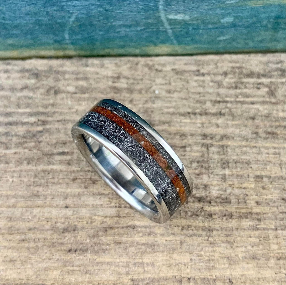 A silver-colored ring with a textured gray band and a reddish-brown stripe, viewed from a slight angle on a wooden surface.