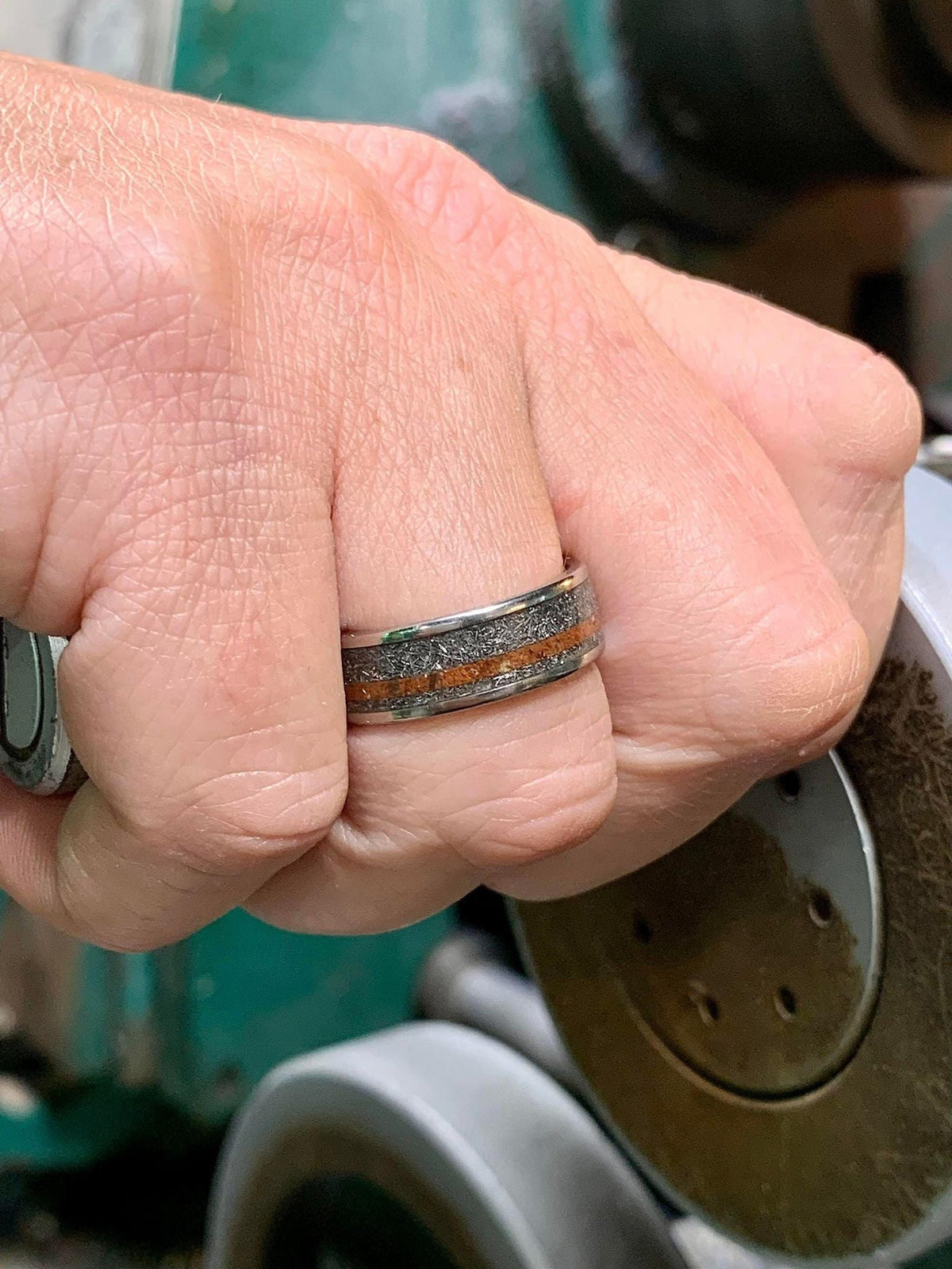 A person's hand wearing a silver-colored ring with a textured gray band and a reddish-brown stripe, holding a metal object.