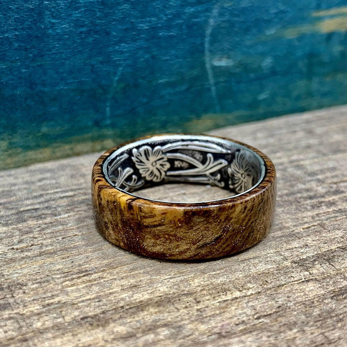 A wooden ring with a silver-colored interior band, featuring an engraved floral pattern, is shown from a slightly elevated angle on a wooden surface.