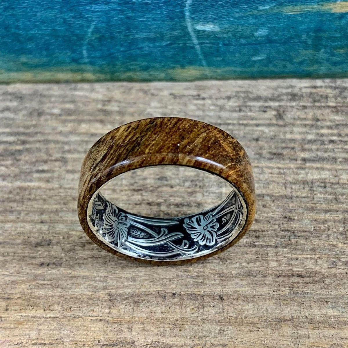 A wooden ring with a silver-colored interior band, engraved with a floral design, is centered on a wooden surface with a blue background.