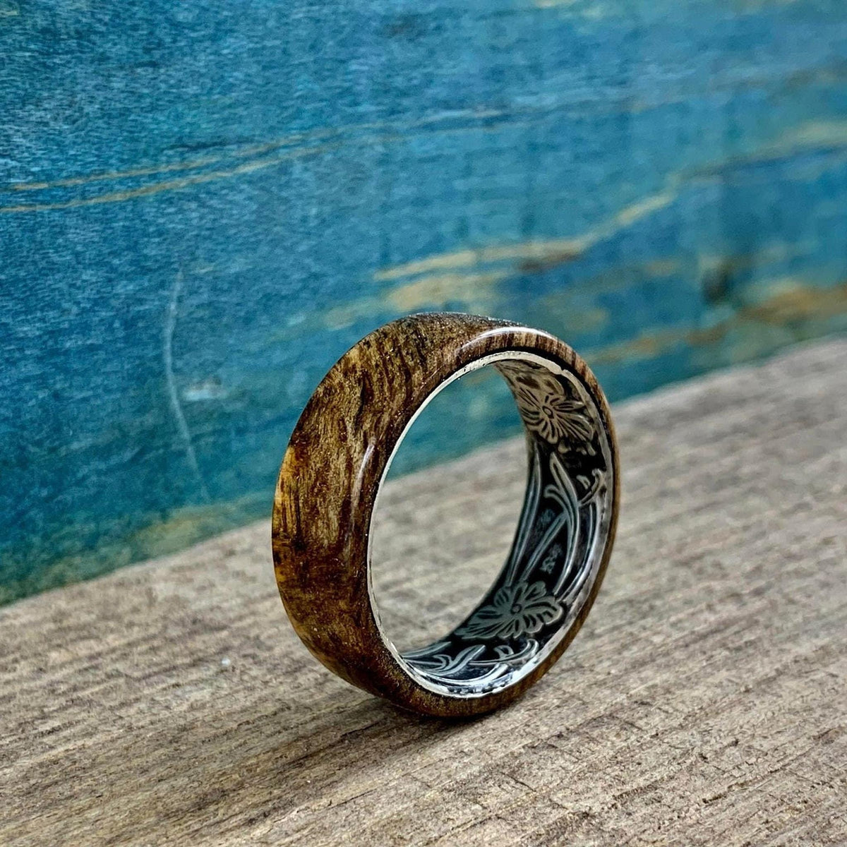 A wooden ring with a silver-colored interior band, engraved with a floral pattern, is displayed at an angle on a wooden surface.