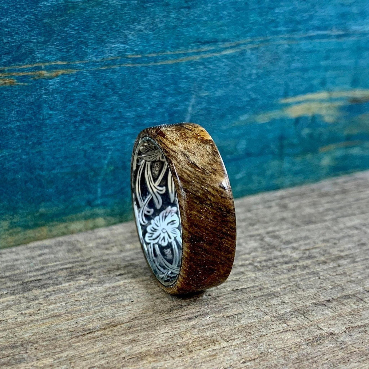 A wooden ring with a silver-colored interior band, engraved with a floral pattern, is shown from a side angle on a wooden surface.