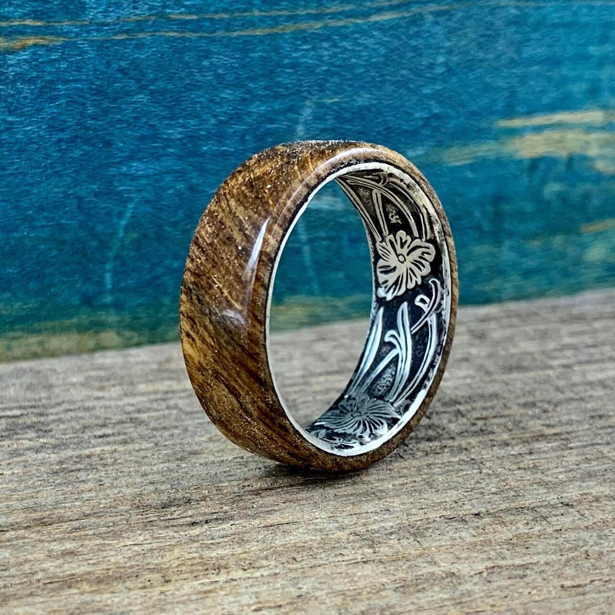 A wooden ring with a silver-colored interior band, engraved with a floral pattern, is shown from a side angle on a wooden surface.