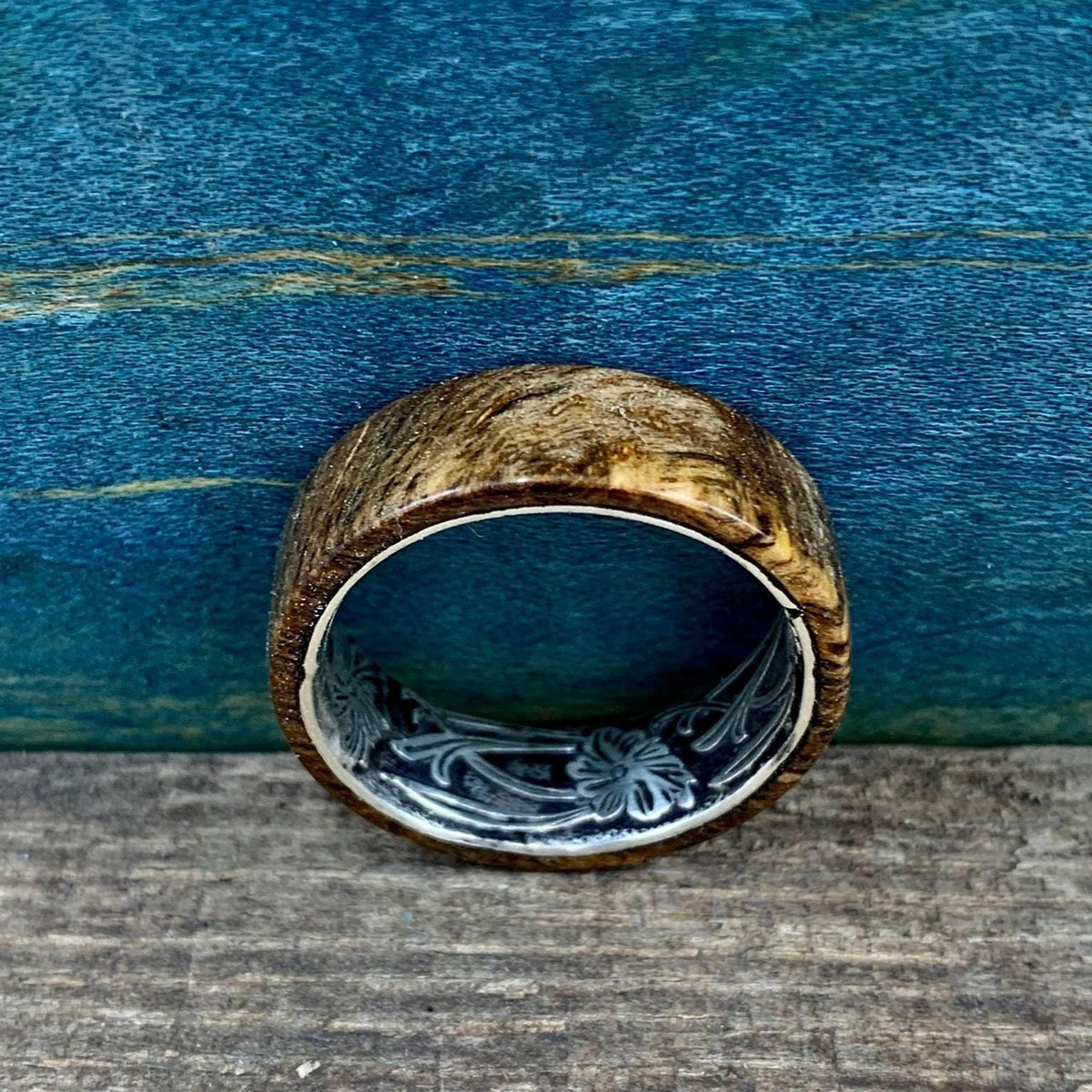 A wooden ring with a silver-colored interior band, engraved with a floral pattern, is shown from a top-down perspective on a wooden surface.