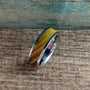 A silver-colored ring with a wide, golden-brown wood inlay is angled on a rustic wooden surface, with a blue-green background.