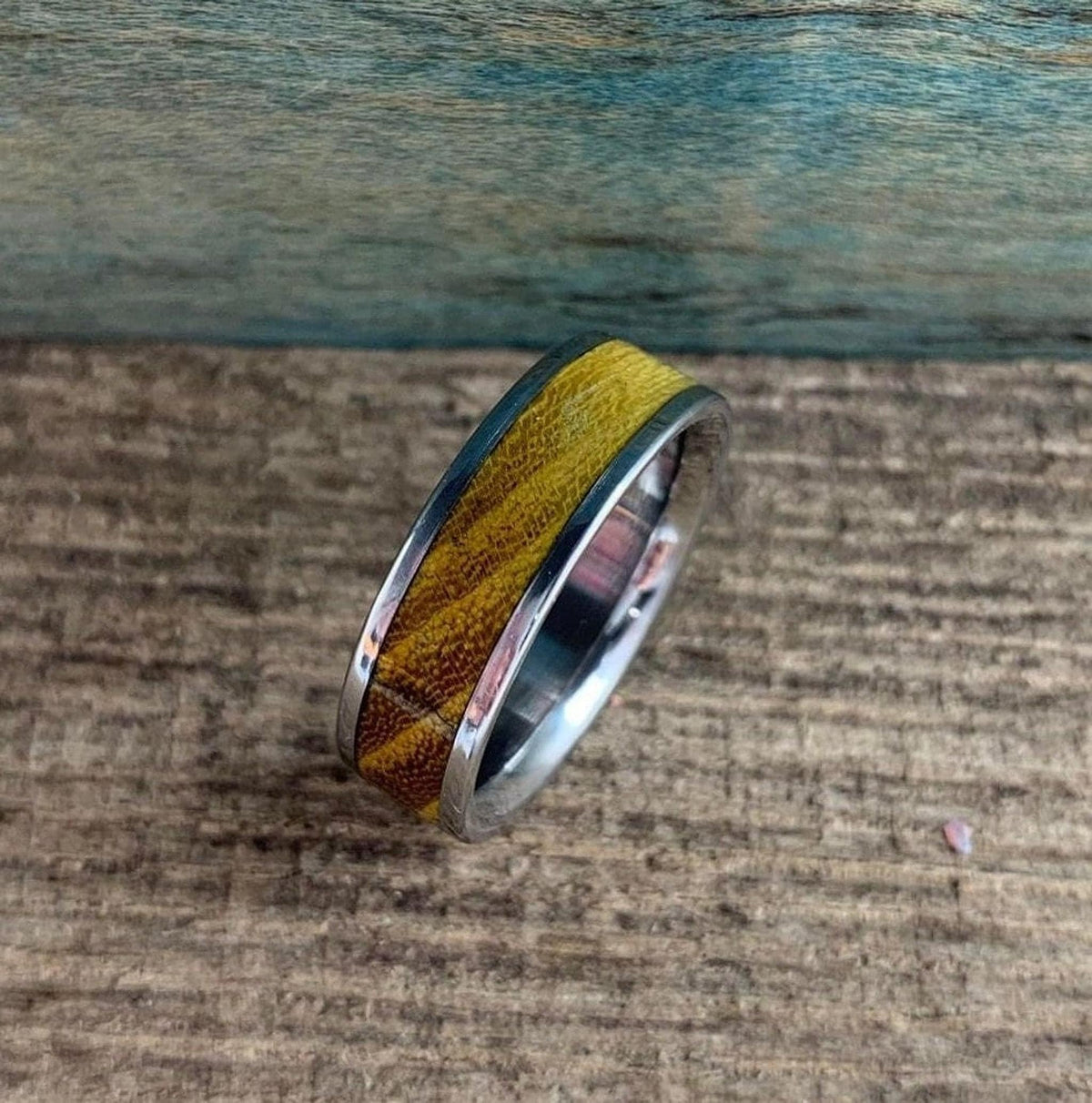 A silver-colored ring with a wide, golden-brown wood inlay is angled on a rustic wooden surface, with a blue-green background.