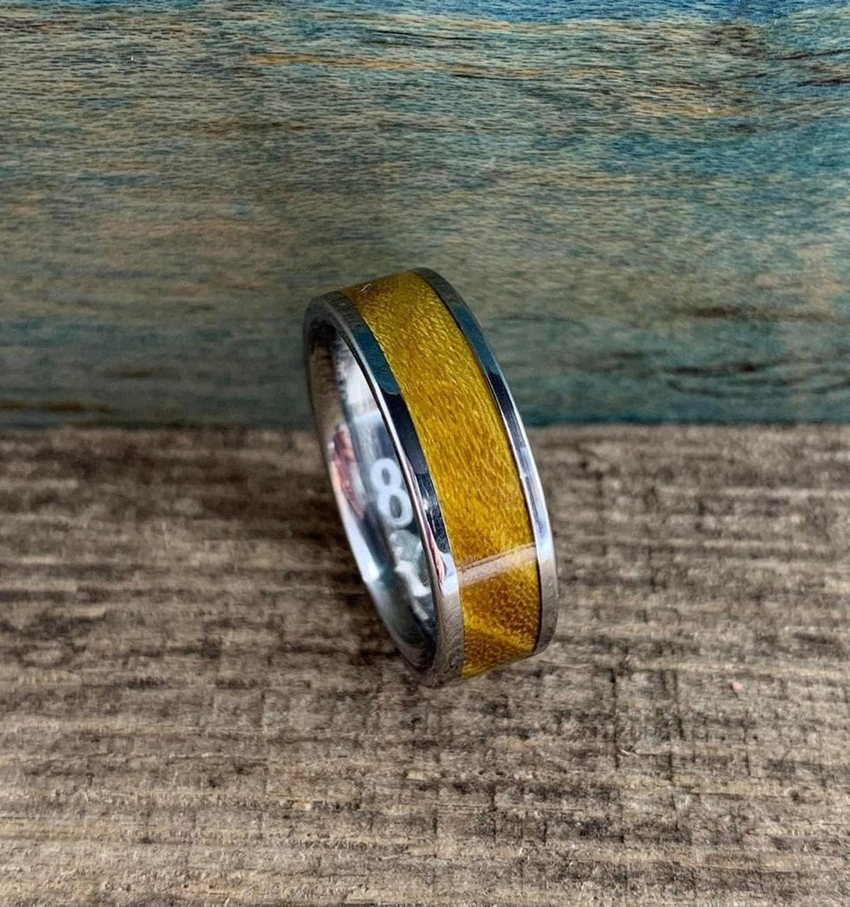 A silver-colored ring with a golden-brown wood inlay is angled, revealing the number "8" engraved on its interior band.
