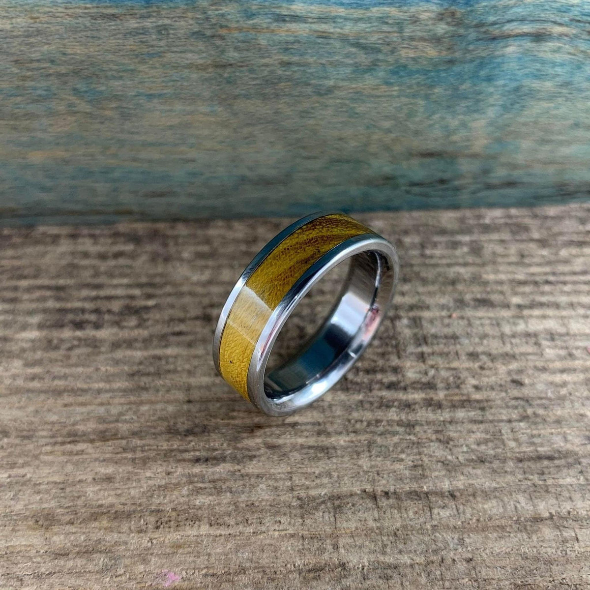 A silver-colored ring with a golden-brown wood inlay is angled on a rustic wooden surface, with a blue-green background.