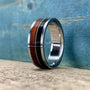 A silver ring with a central band of dark wood, a thin silver guitar string, and a band of reddish-brown wood, sits on a light wooden surface against a blue background.