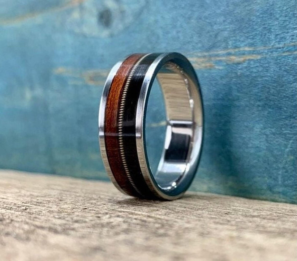 A silver ring with a central band of dark wood, a thin silver guitar string, and a band of reddish-brown wood, sits on a light wooden surface against a blue background.