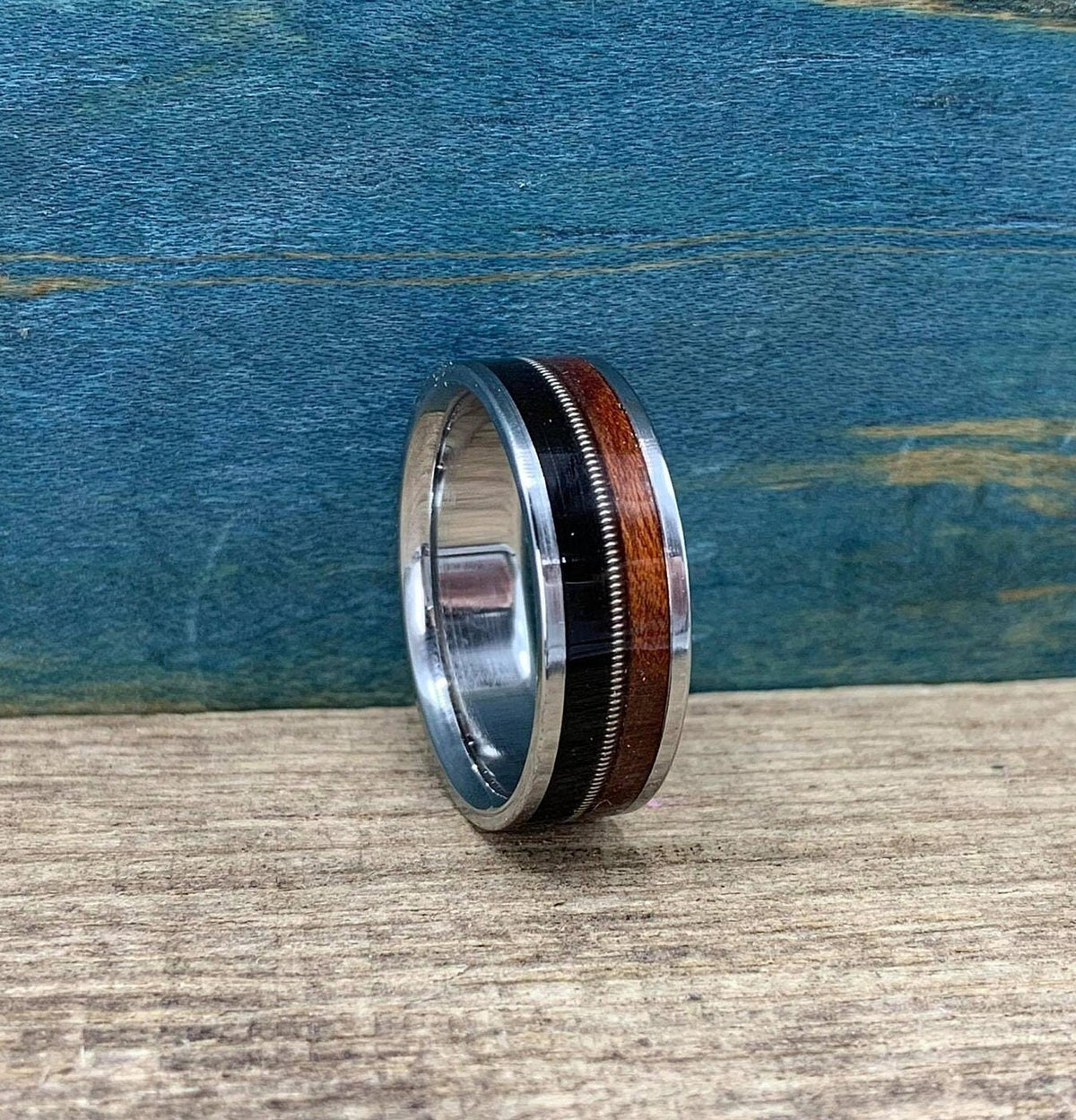 A silver ring with a central band of dark wood, a thin silver guitar string, and a band of reddish-brown wood, is shown from a slightly different side angle on a light wooden surface.