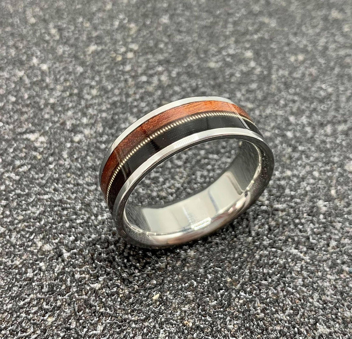 A silver ring with a central band of dark wood, a thin silver guitar string, and a band of reddish-brown wood, is displayed on a dark, textured surface.