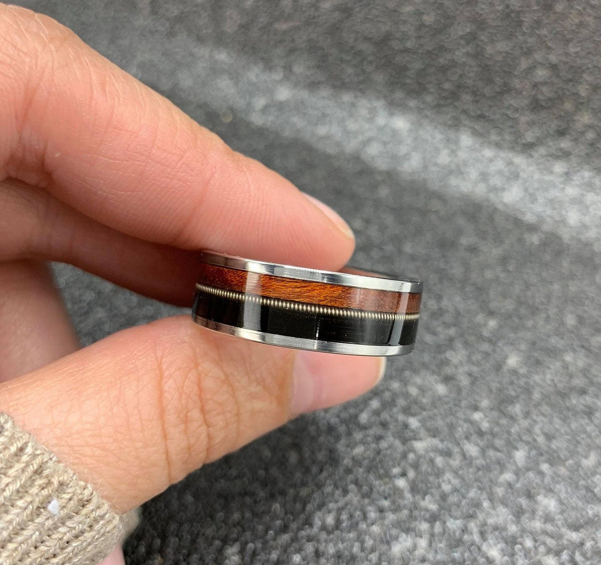 A hand holds a silver ring with a central band of dark wood, a thin silver guitar string, and a band of reddish-brown wood, against a dark, textured background.