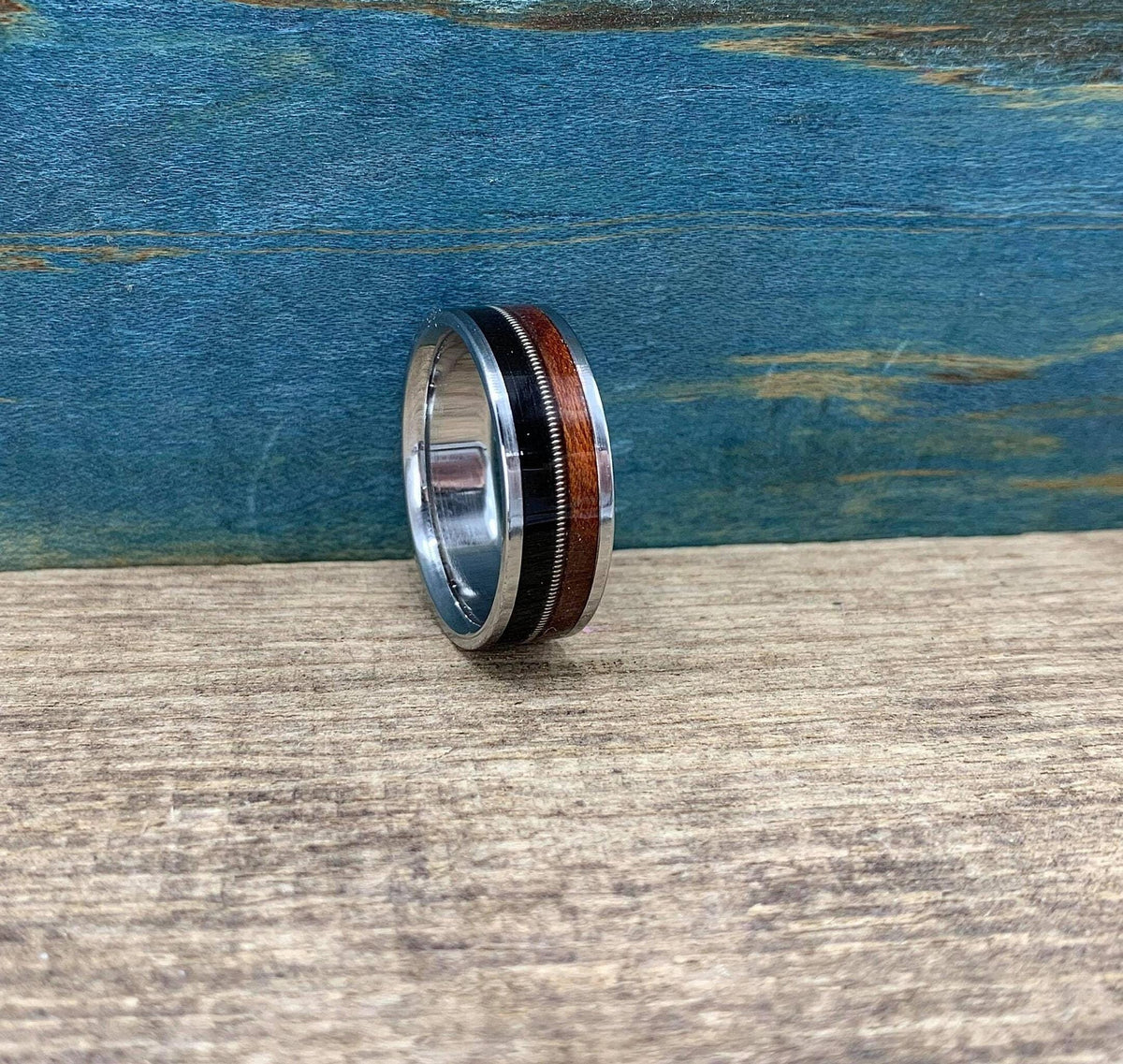 A silver ring with a central band of dark wood, a thin silver guitar string, and a band of reddish-brown wood, is shown from a front-facing angle on a light wooden surface.