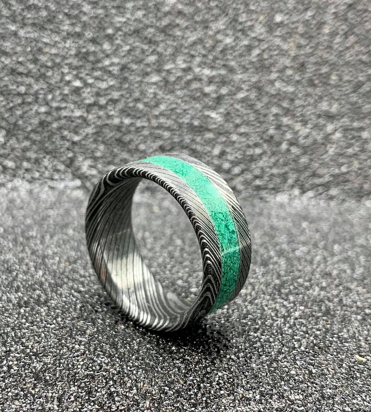 A Damascus steel ring with a malachite inlay, displayed on a dark, textured surface.