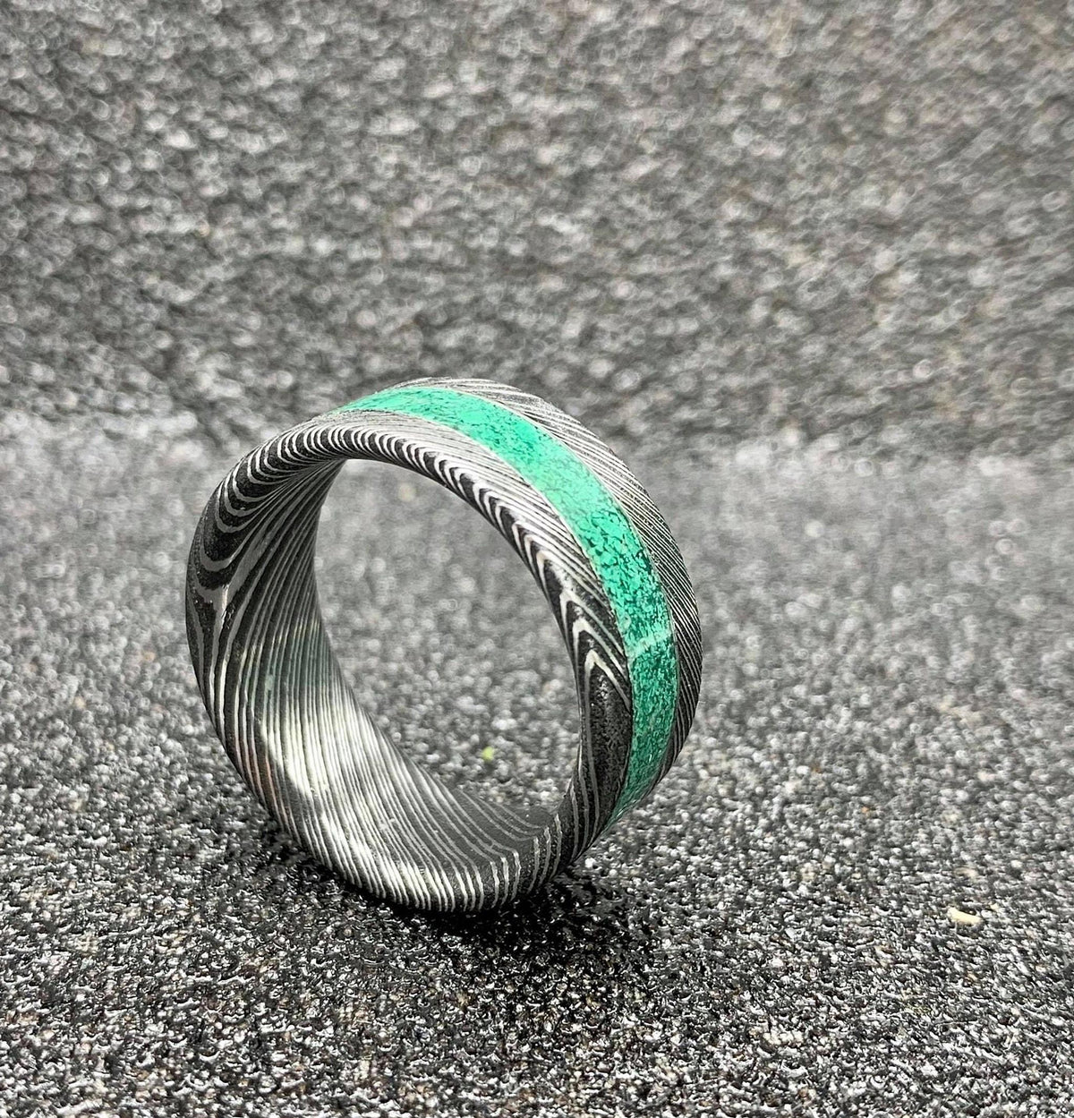 A Damascus steel ring with a malachite inlay, shown from the side on a dark, textured surface.