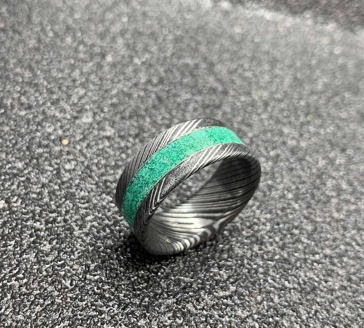 A Damascus steel ring with a malachite inlay, presented on a dark, textured surface.