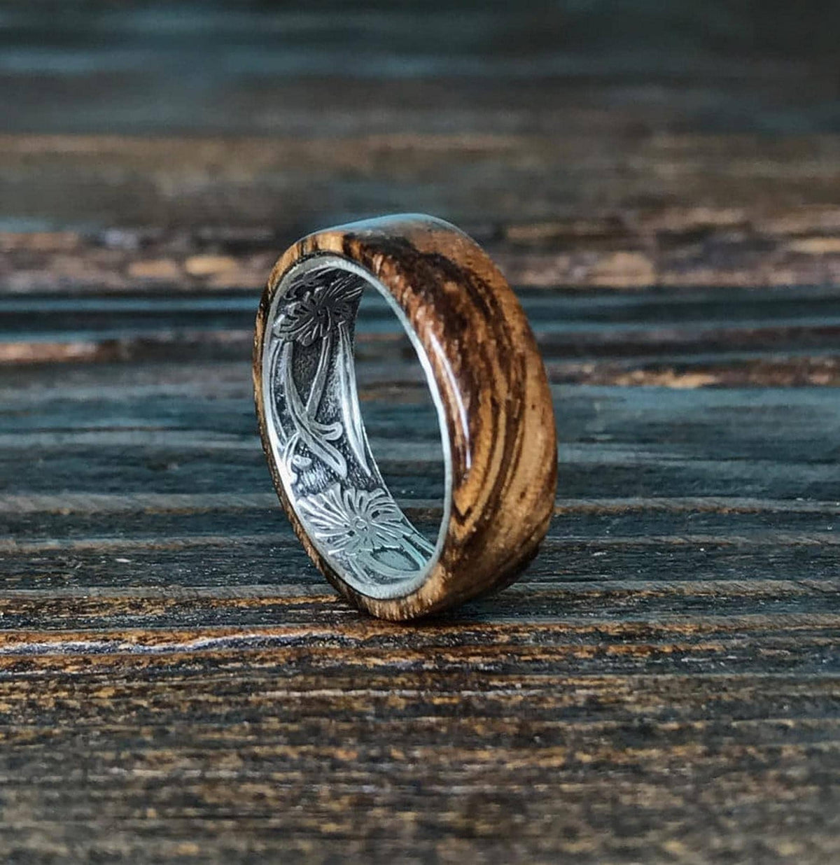 A zebrawood ring with a silver interior and engraved floral pattern is angled on a dark wooden surface, showcasing the wood grain.