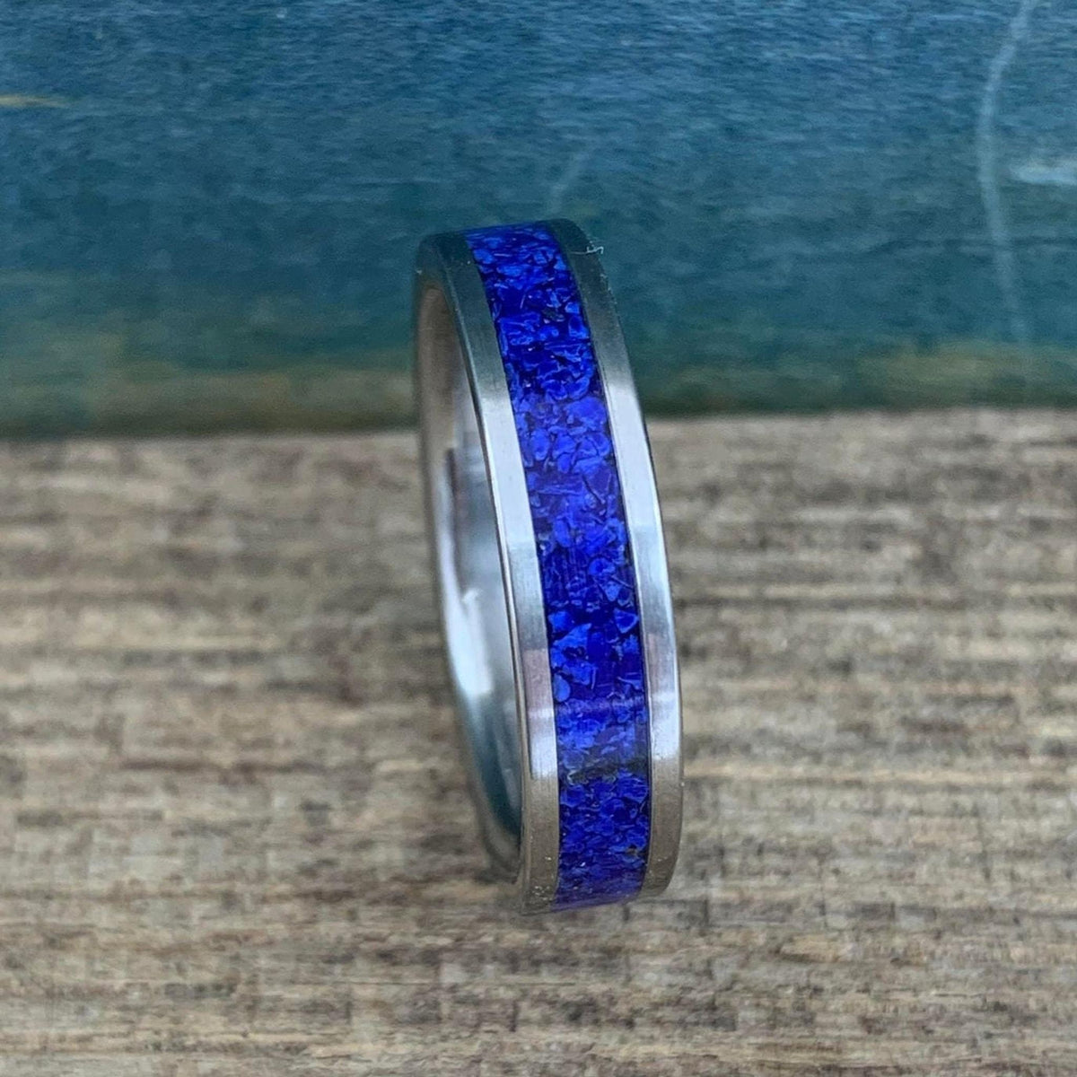 A silver-colored titanium ring with a central band of crushed blue lapis lazuli, viewed from a slight angle on a wooden surface.