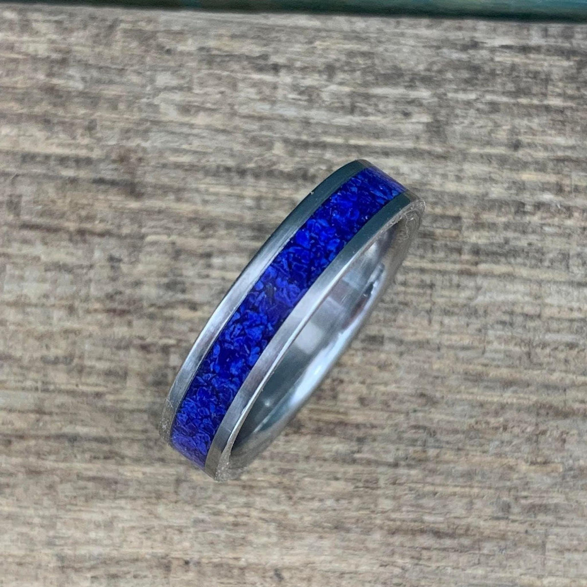 A silver-colored titanium ring with a central inlay of crushed blue lapis lazuli, viewed from above on a light wooden surface.