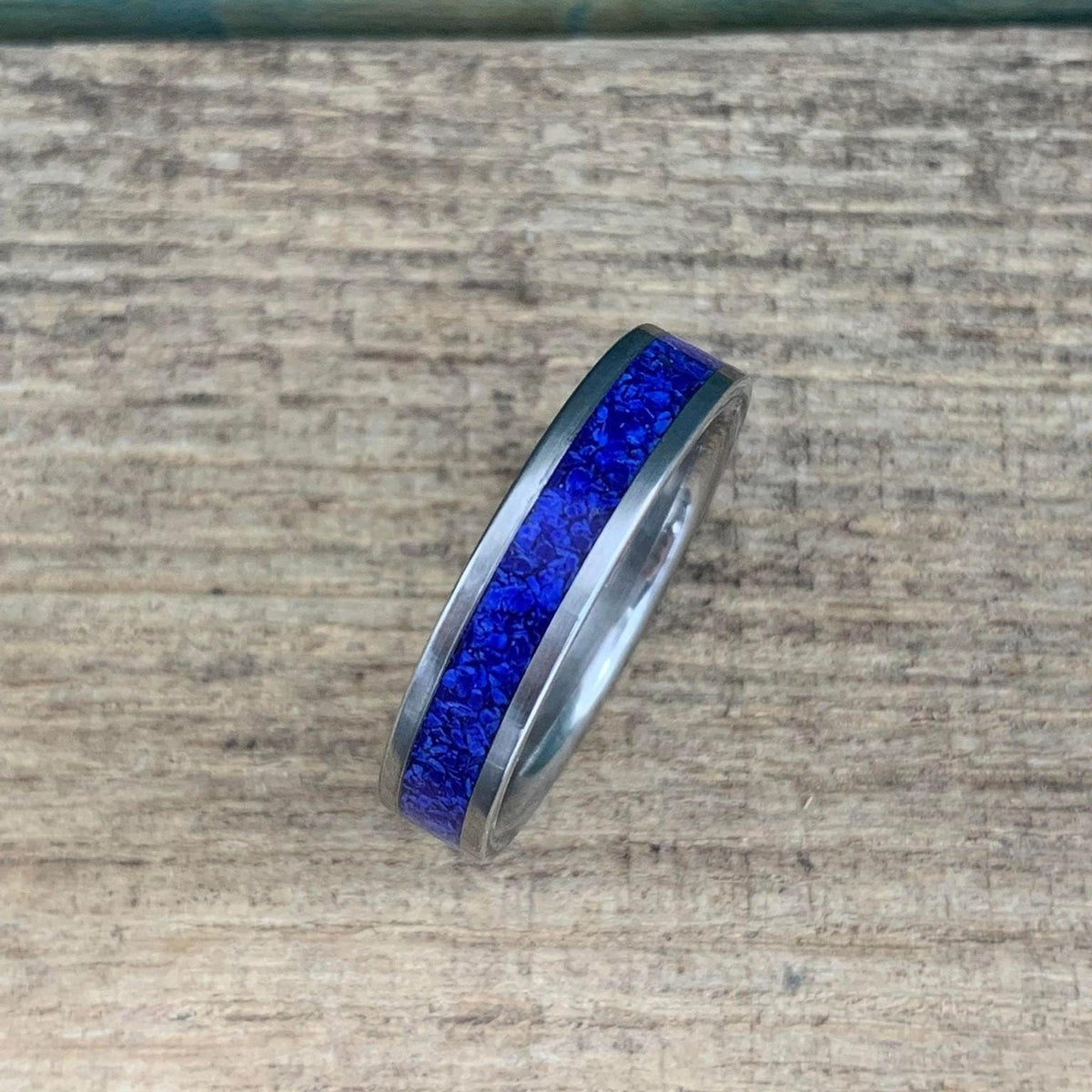 A silver-colored titanium ring with a central band of crushed blue lapis lazuli, viewed from above on a light wooden surface.