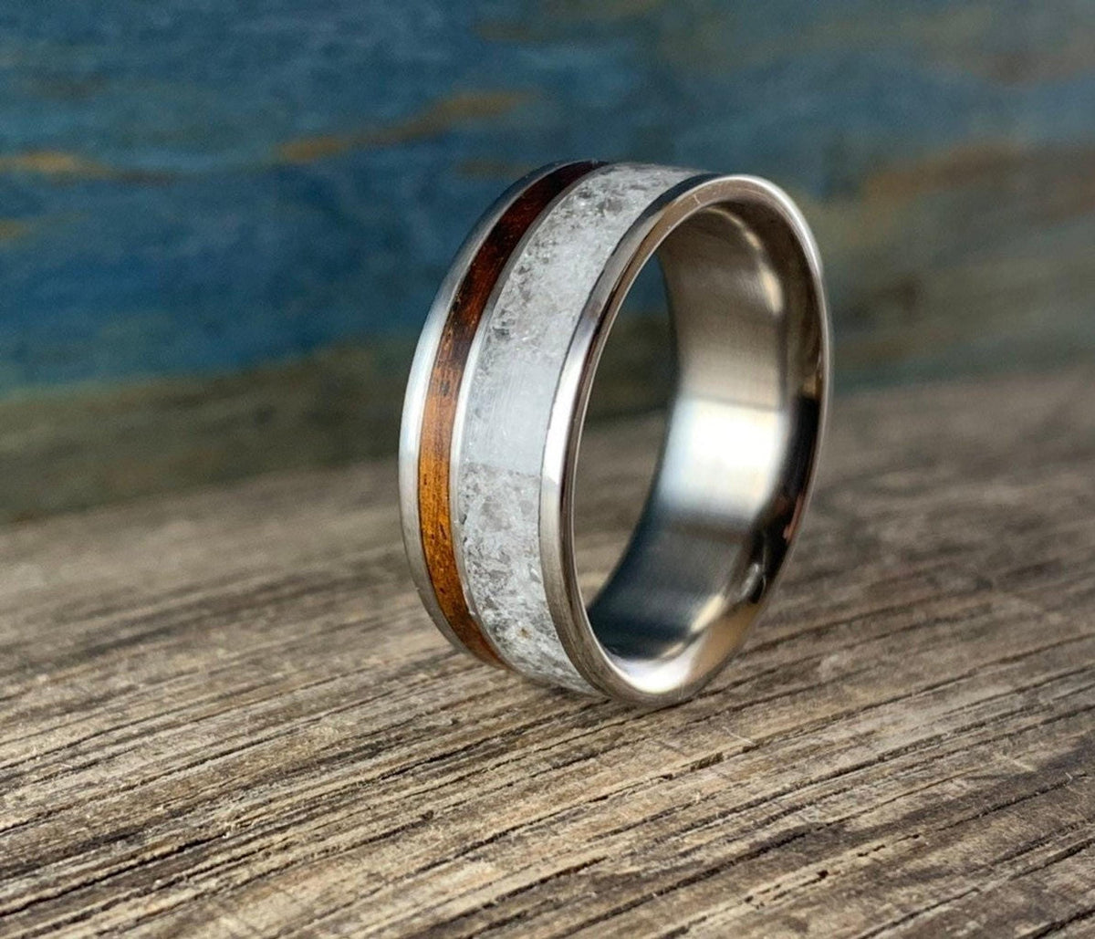 A titanium ring with a central band of white rose quartz and a narrow strip of dark kingwood, shown at an angle on a wooden surface.