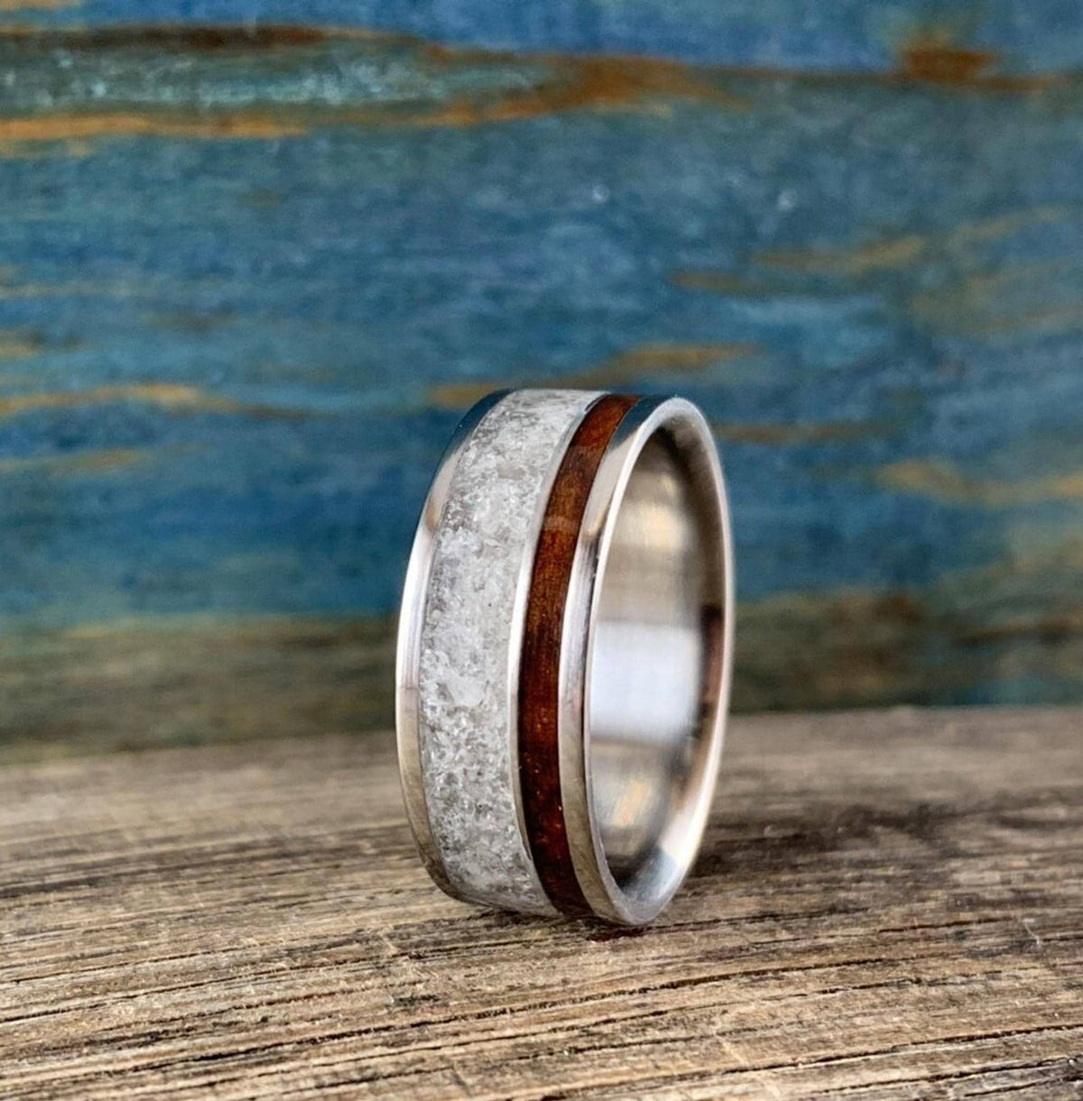 A titanium ring with a central band of white rose quartz and a narrow strip of dark kingwood, shown at an angle on a wooden surface.