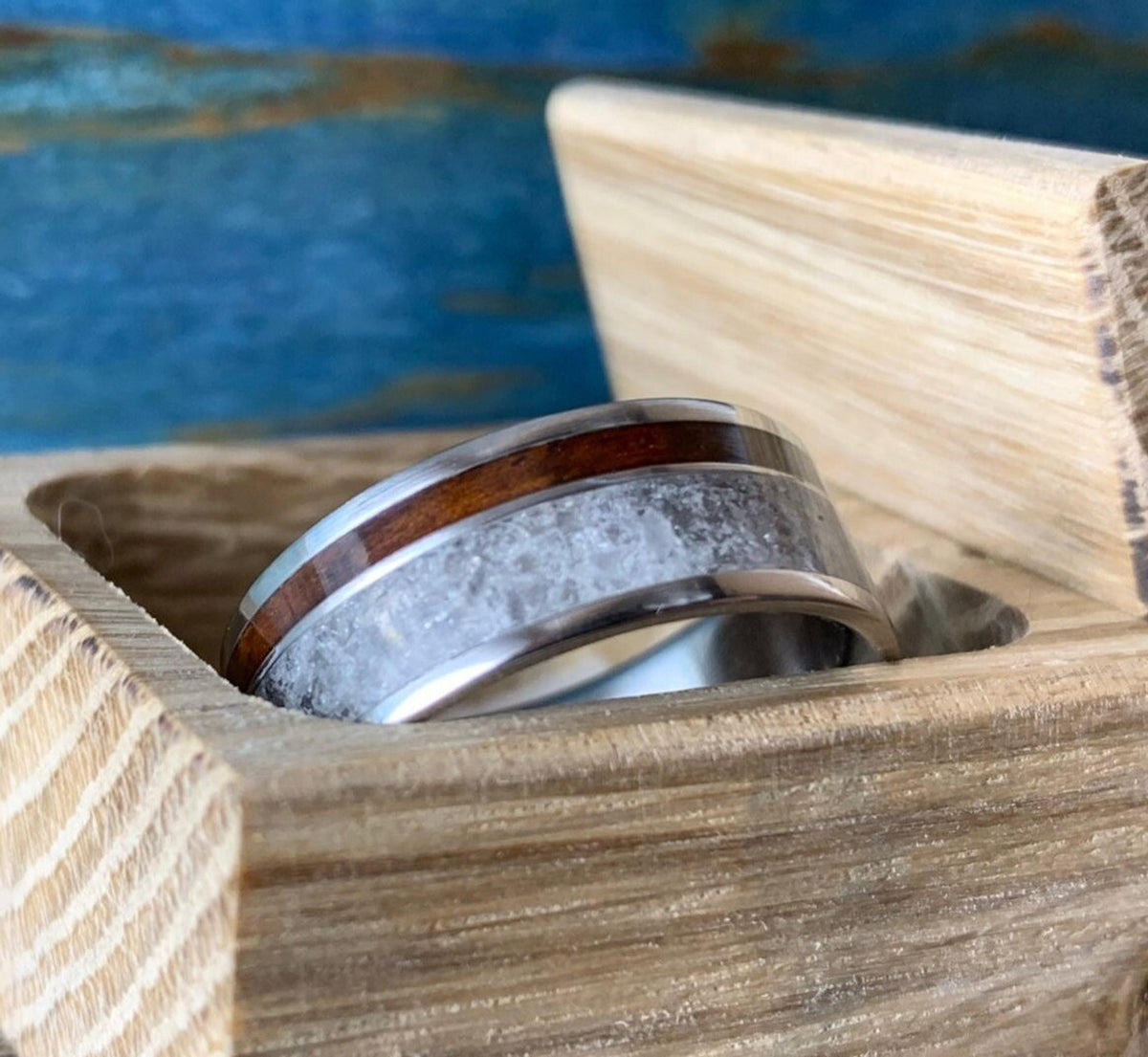 A titanium ring with a central band of white rose quartz and a narrow strip of dark kingwood, resting inside a light wooden box.