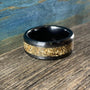 A black ceramic ring with a textured gold-colored inlay is shown at an angle on a wooden surface with a blue background.