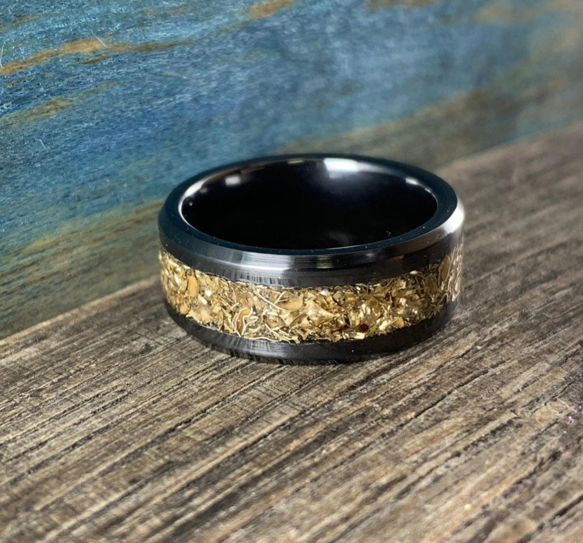 A black ceramic ring with a textured gold-colored inlay is shown at an angle on a wooden surface with a blue background.