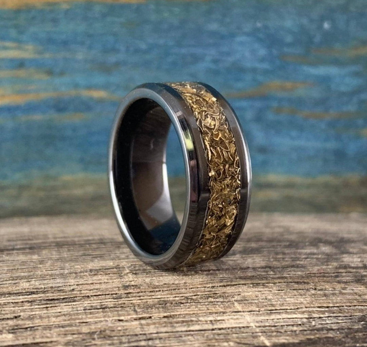 A black ceramic ring with a textured gold-colored inlay is shown from the side on a wooden surface with a blue background.