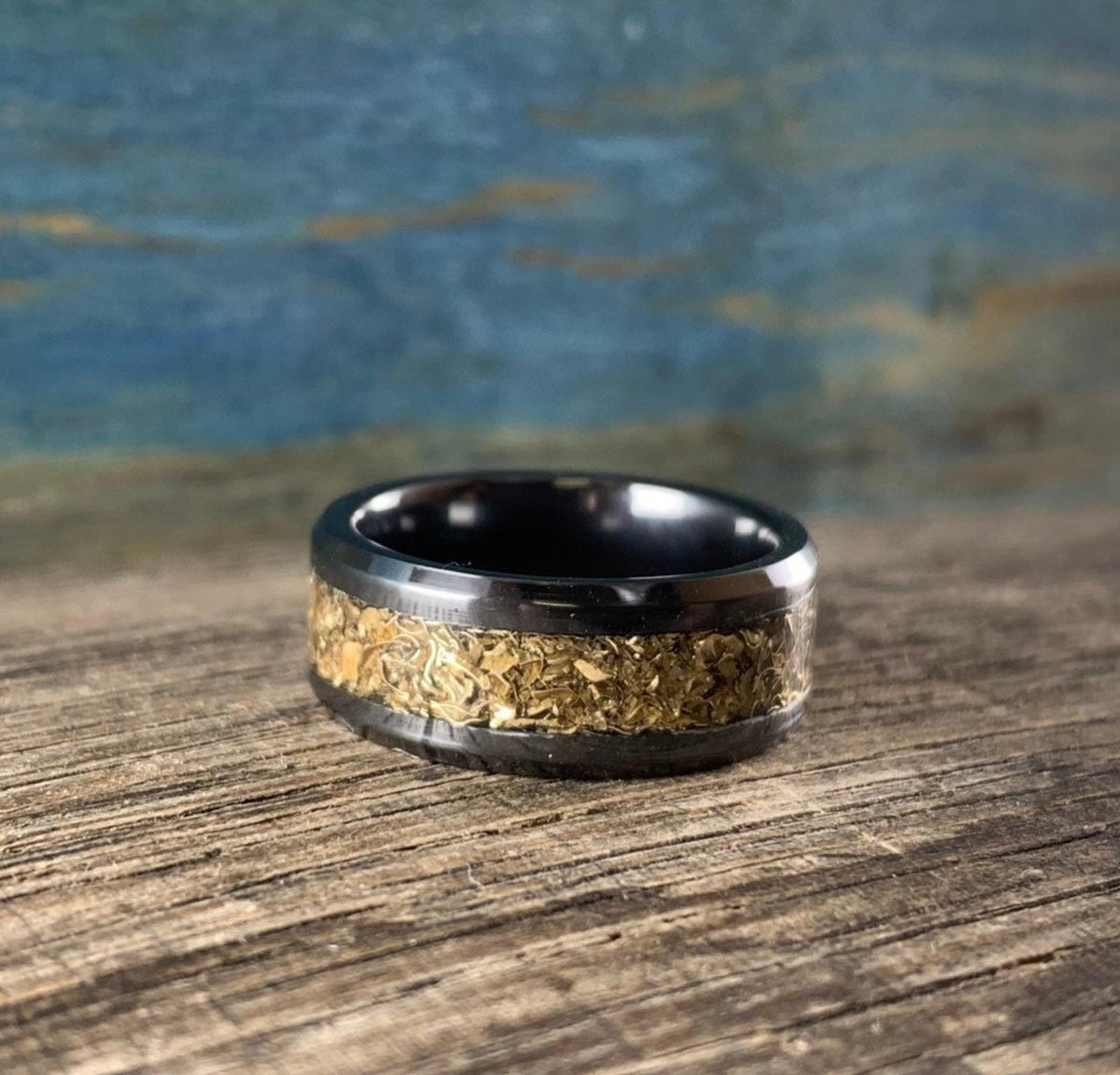 A black ceramic ring with a textured gold-colored inlay is shown from the front on a wooden surface with a blue background.