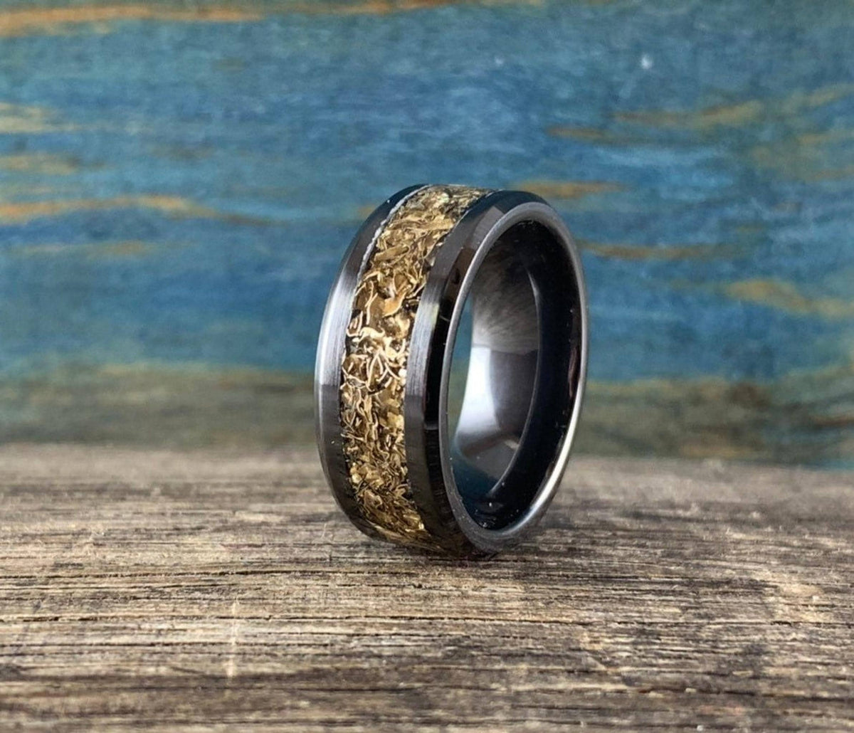 A black ceramic ring with a textured gold-colored inlay is shown from the side on a wooden surface with a blue background.