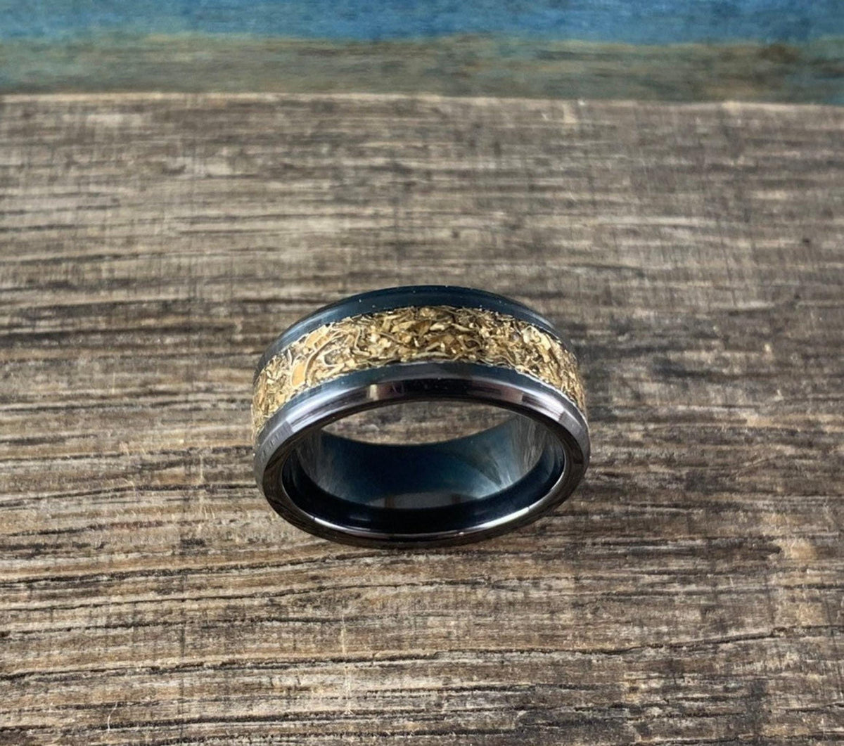 A black ceramic ring with a textured gold-colored inlay is shown from above on a wooden surface with a blue background.