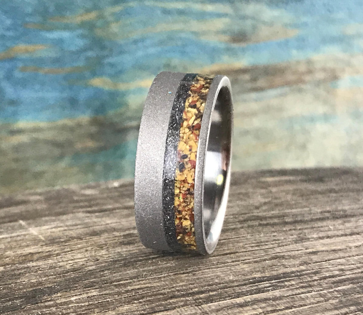 A silver-colored ring with a sandblasted finish and a central band of yellow, orange, and black flecks, stands upright on a wooden surface.
