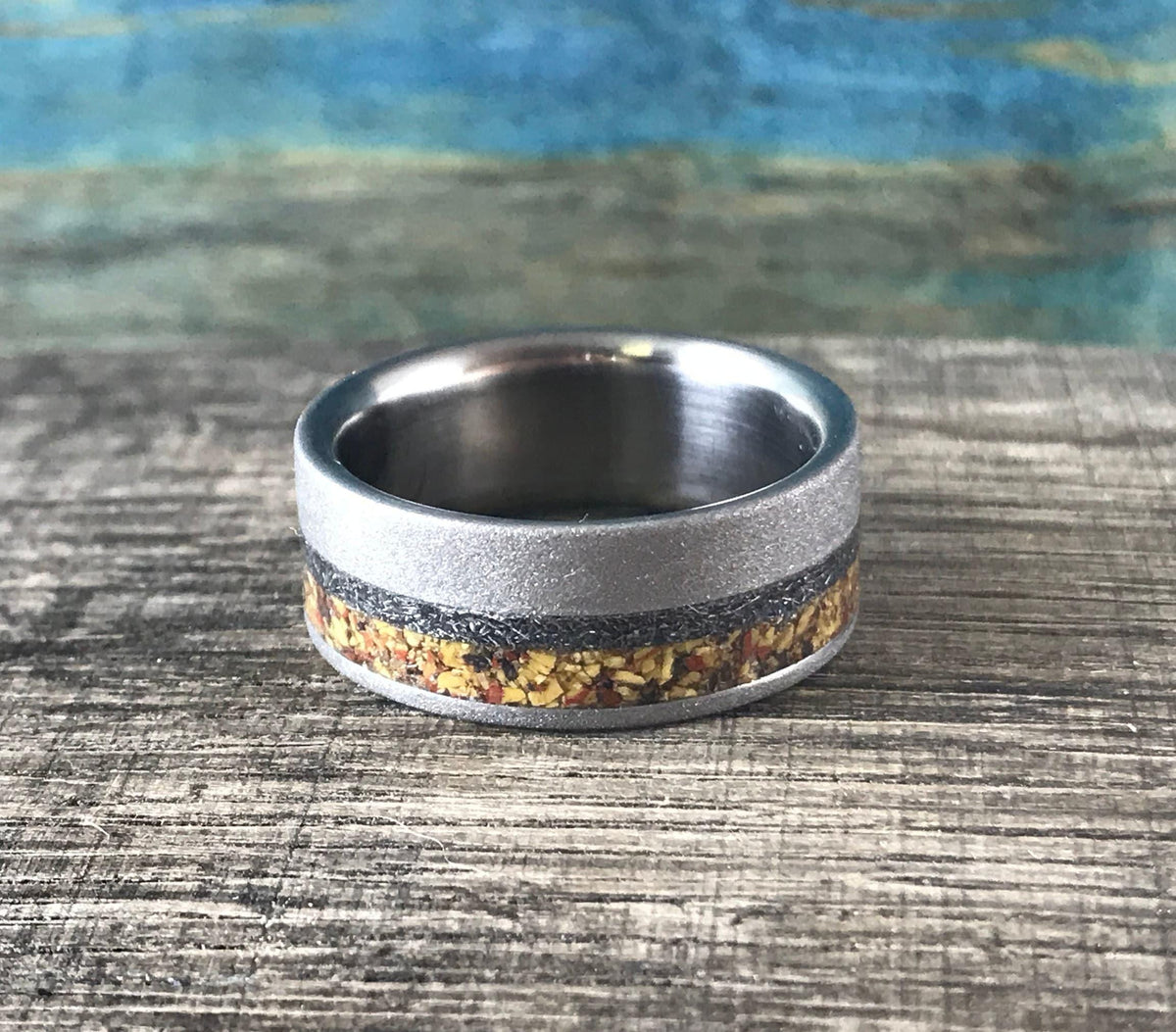 A silver-colored ring with a sandblasted finish and a central band of yellow, orange, and black flecks, is viewed from the side on a wooden surface.