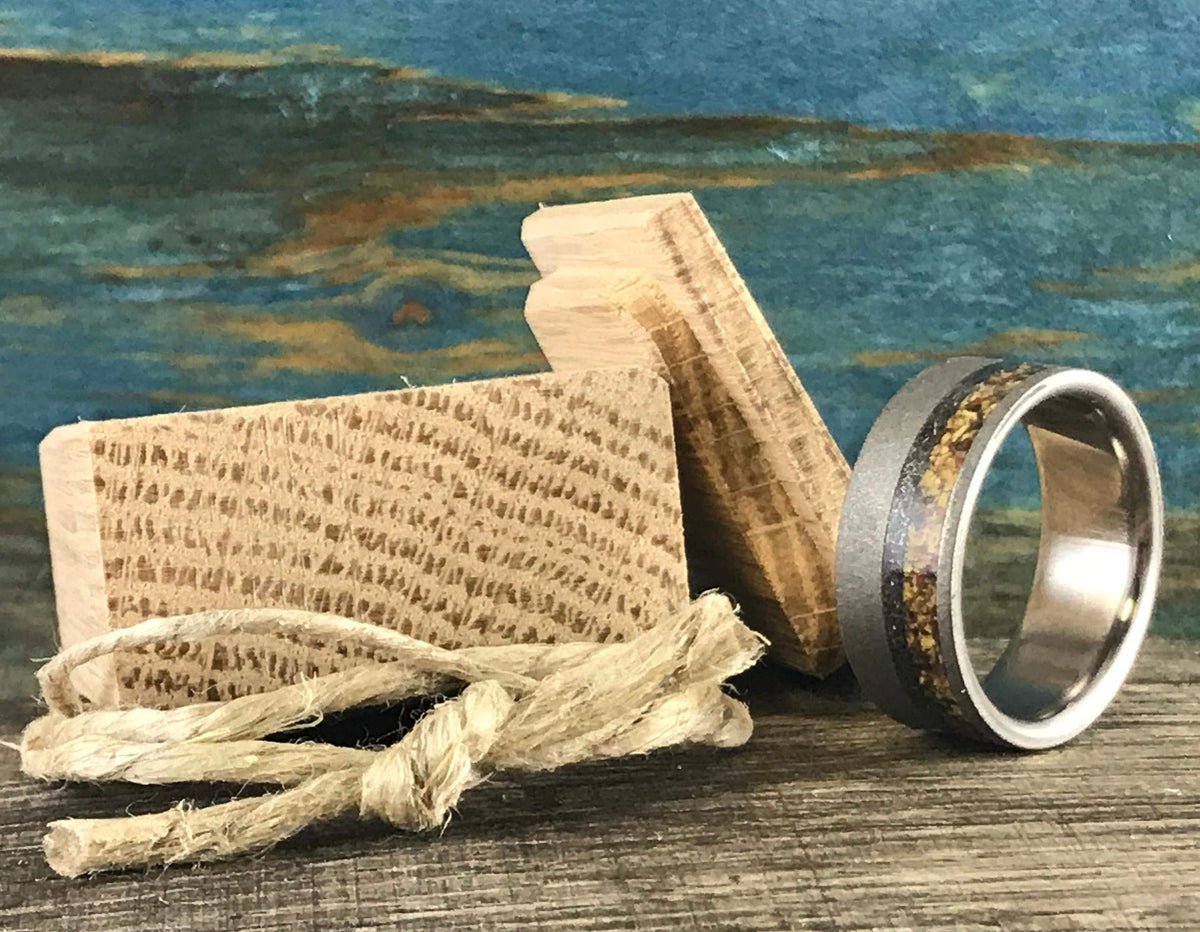 A silver-colored ring with a sandblasted finish and a central band of yellow, orange, and black flecks, sits next to two small wooden blocks and a rope.