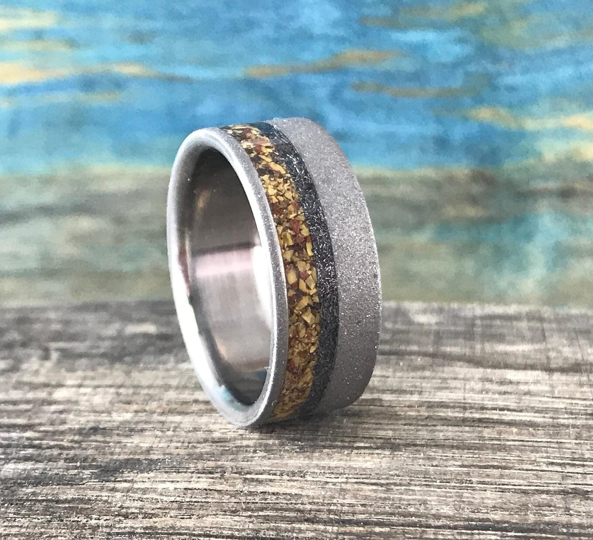 A silver-colored ring with a sandblasted finish and a central band of yellow, orange, and black flecks, is angled slightly on a wooden surface.