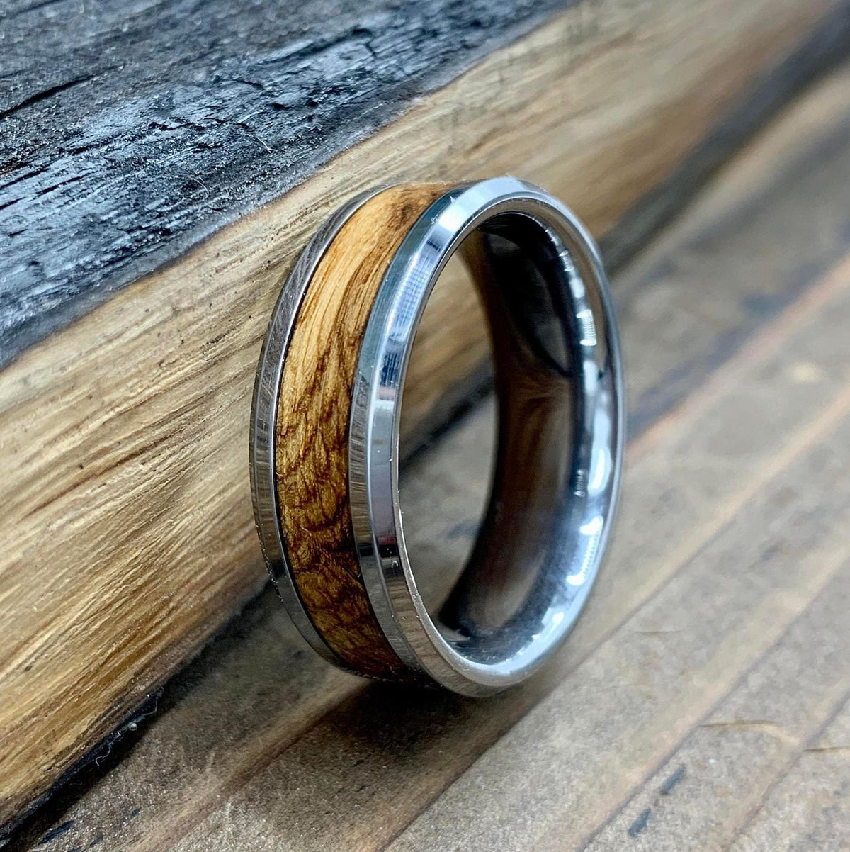 A silver-colored ring with a central light brown wood inlay, resting on a rustic wooden plank.